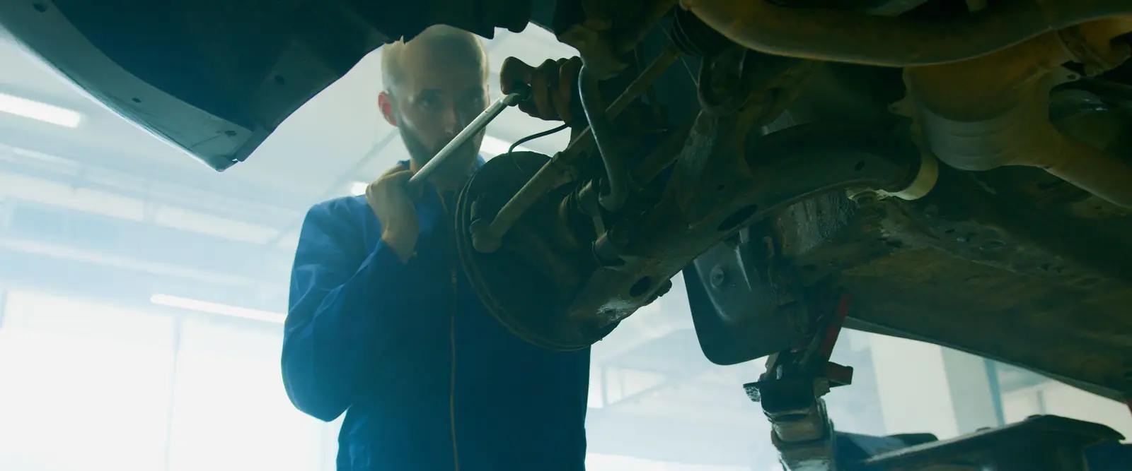 A mechanic in blue coveralls stands under a car raised on a lift and turns a wrench on the front suspension in a hazy, brightly lit workshop.