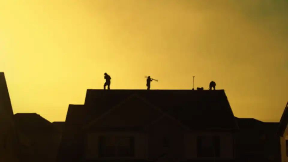 Three roofers are silhouetted on the roof ridge of a house against a yellow evening sky, one carrying a long-handled fork tool on his shoulder.