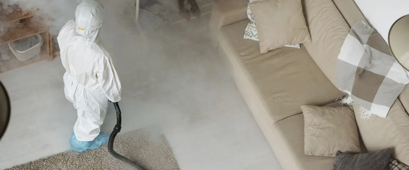 Seen from above, a worker in a white protective suit, hood and blue shoe covers runs a fogging machine in a living room that is filling with mist.