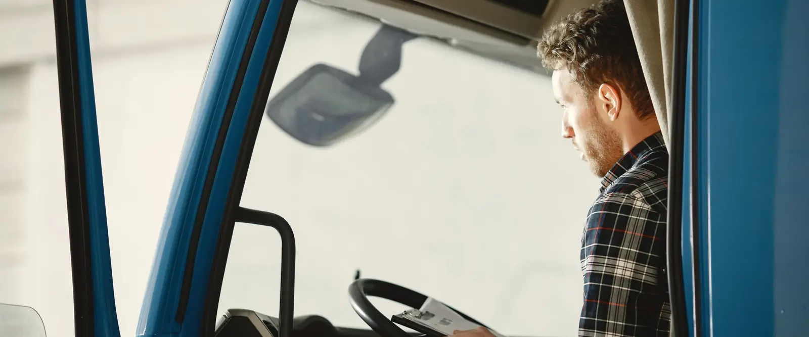 A man in a plaid shirt sits in the cab of a blue truck with the door open and reads papers on a clipboard resting on the steering wheel.